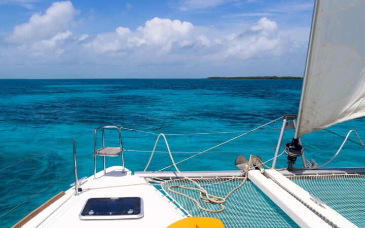 the view from the rear of a Belize catamaran charter.The white netting and white sails sit against a crystal blue and teal ocean horizon and cloudy sky