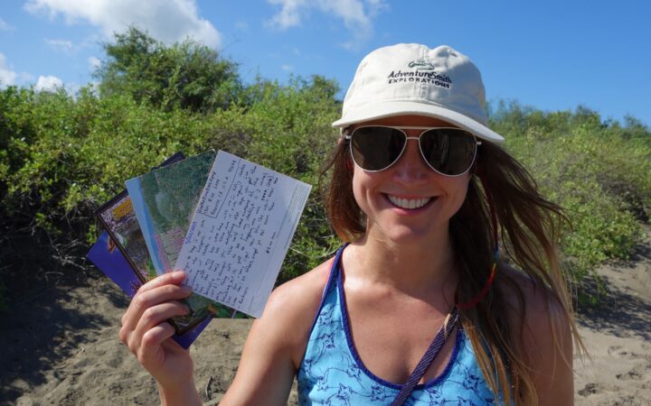 A female traveler with a tan hat and sunglasses holds up postcards taken at Postcard Bay in the Galapagos Islands.