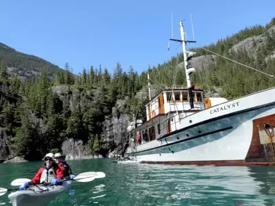 On a blue sunny day in Alaska the small ship Catalyst floats on blue water as two guests paddle a double kayak near by
