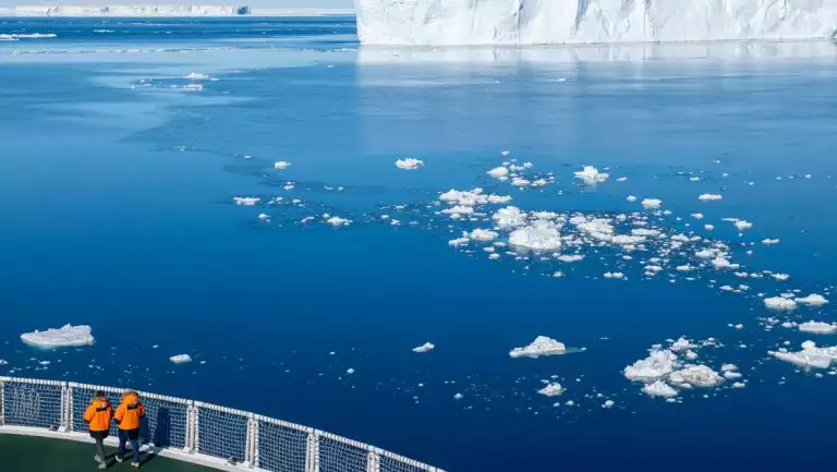 2 travelers in orange coats aboard a Le Commandant Charcot Weddell Sea Voyage stand in sunshine & view a large tabular iceberg.