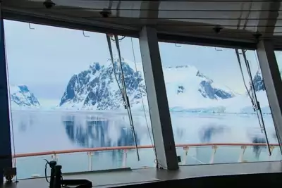 Looking out through glass windows from the observation deck towards a jagged snow covered mountain range in Antarctica