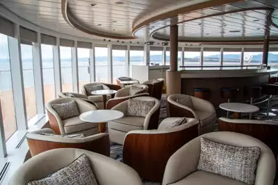 Observation lounge filled with cream upholstered chairs set around small coffee tables in front of wrap around floor to ceiling windows.