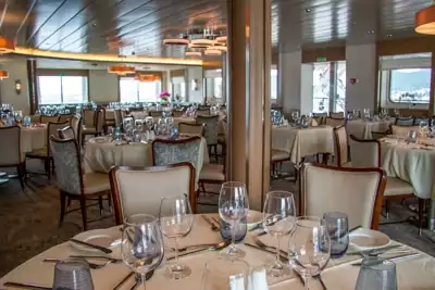 Dining room with round tables set with table settings, glasses and upholstered cream chairs with wood trim aboard Greg Mortimer.