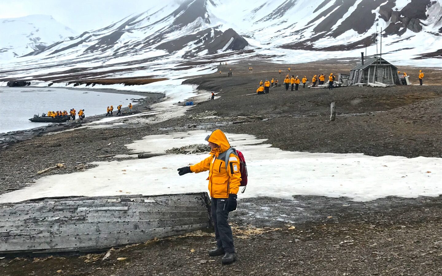 Arctic travelers wearing yellow parkas explore the shores of the Svalbard archipelago on a cloudy day surrounded by snowy mountain range.