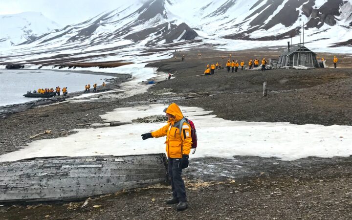 Arctic travelers wearing yellow parkas explore the shores of the Svalbard archipelago on a cloudy day surrounded by snowy mountain range.