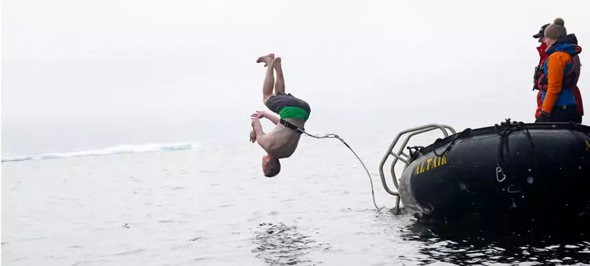 Polar plunge! A man flips into the arctic ocean in front of an ice shelf. He is harnessed to a black inflatable skiff for safety.