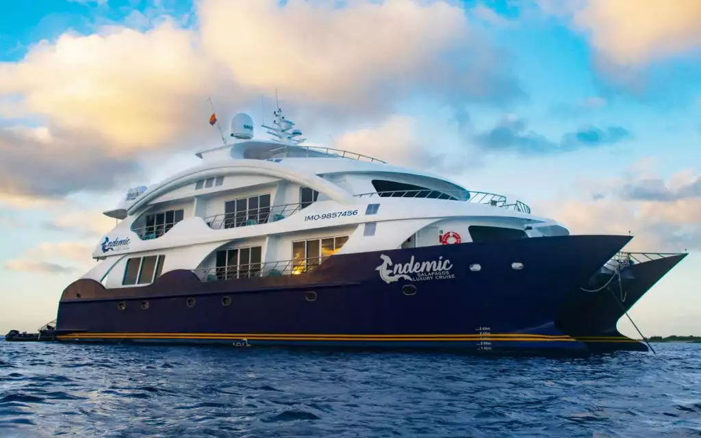 Exterior of Endemic catamaran sitting in the water, with twin dark blue hulls & 2 white upper decks, on a partly cloudy Galapagos day.