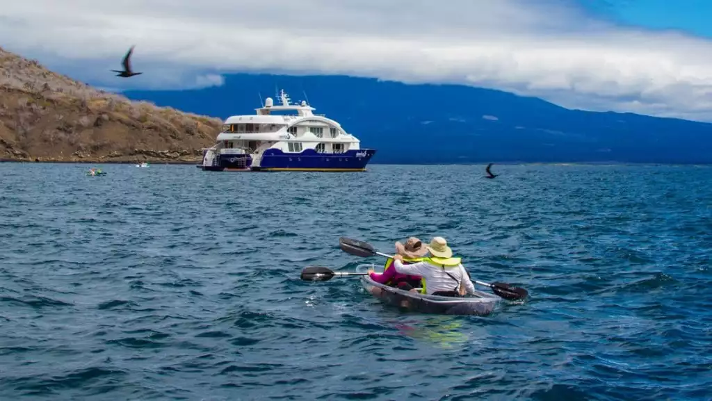 Tandem kayakers in bright yellow PFDs paddle calm water toward a blue & white catamaran with birds overhead on a sunny day in the Galapagos.