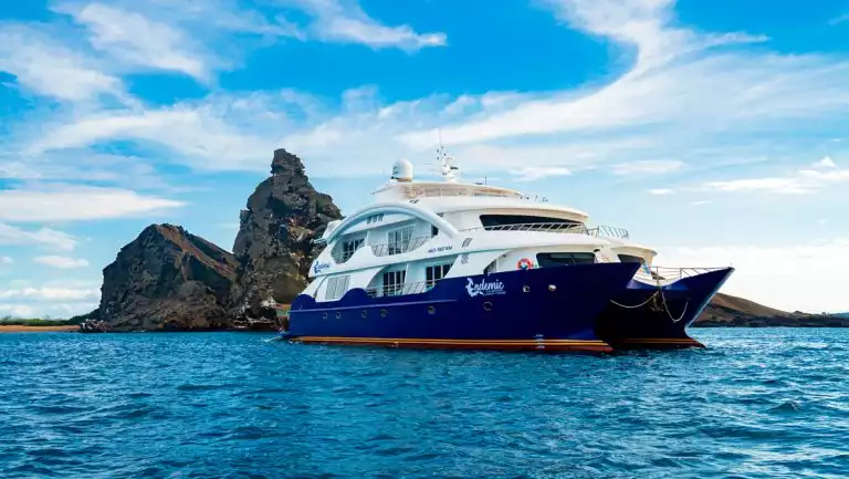 Endemic Galapagos catamaran with blue hull & 2 white upper decks sits in calm sea beside jagged dark rocks under sunny skies.