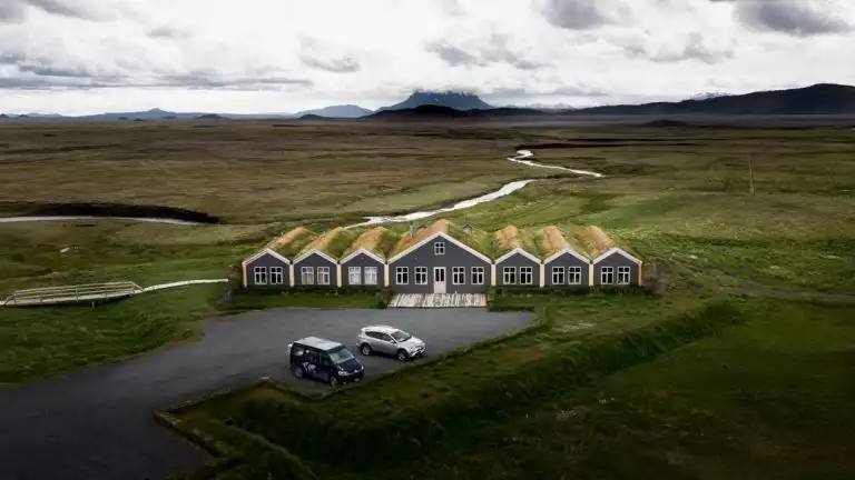 Aerial view of 6 slender grey buildings, 3 on each side of a wider one, with turf roofs & parking lot with 2 cars out front.