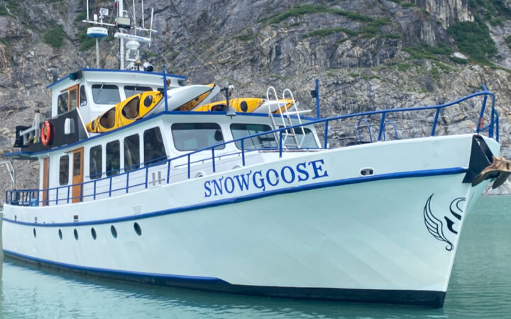 Exterior of Snow Goose Alaska yacht sitting in calm water beside rocky cliffs, with white walls, blue hull & 3 decks.
