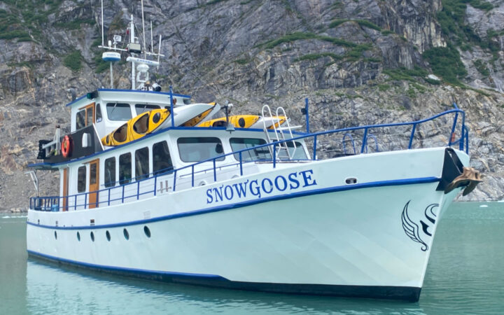 Exterior of Snow Goose Alaska yacht sitting in calm water beside rocky cliffs, with white walls, blue hull & 3 decks.