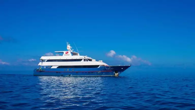 Aerial view of Calipso yacht in Galapagos with dark blue hull & white upper decks, cruising through calm blue water on a sunny day.