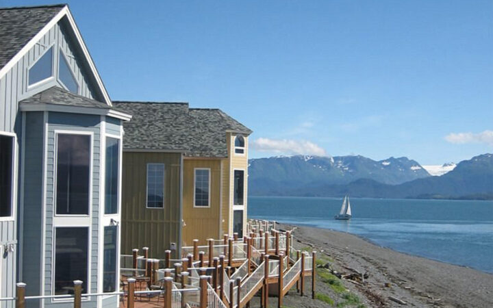 Exterior of blue & yellow Land's End Resort guest rooms sitting seaside with raised platform decks overlooking a sailboat.