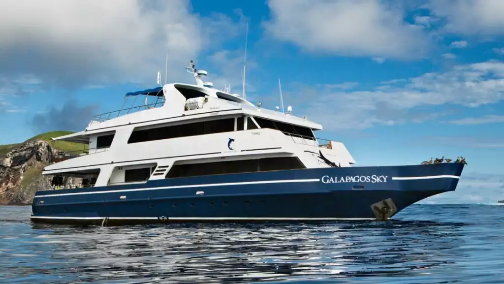 Water-level view of Galapagos Sky dive boat with blue hull & 2 white upper decks, sitting in calm water beside a green island.