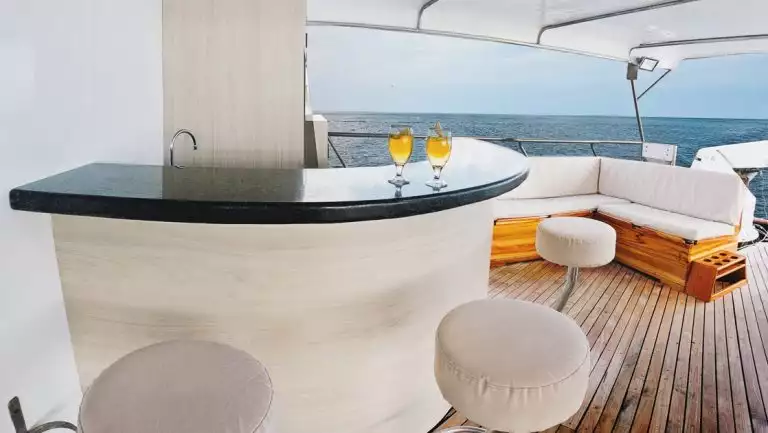 Bacl & white outdoor bar with 3 padded white bar stools & nearby corner couch with teak decking under shade on mv Galapagos Sky.