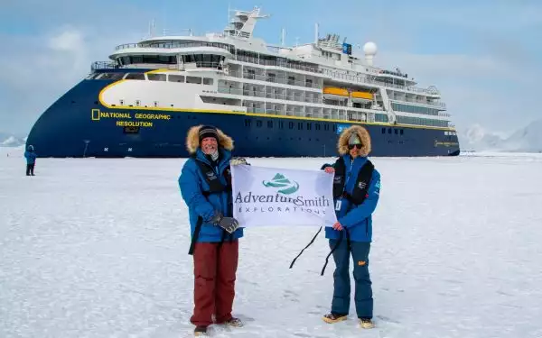 Two male travelers in blue parkas with fur lined hoods, hold an AdventureSmith flag in front of the Nat Geo Resolution ship.