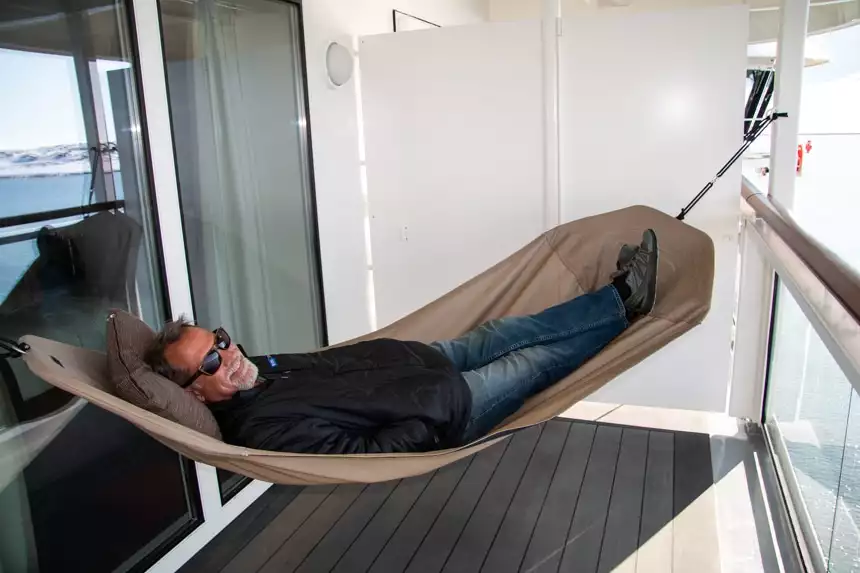 Guest wearing black jacket and blue jeans lays in a hammock strung up on the cabin balcony aboard Nat Geo Resolution.