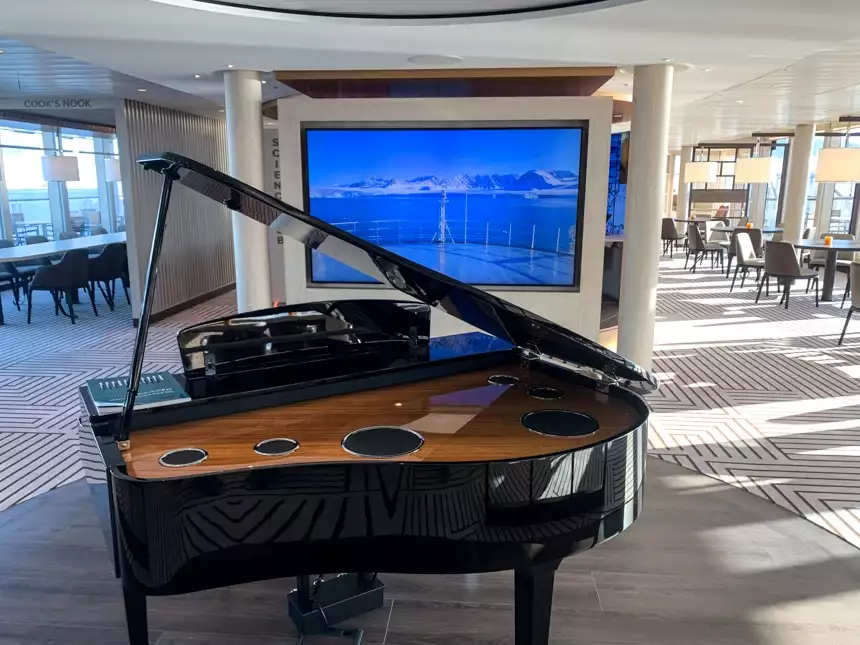 Observation Deck with seating in front of floor-to-ceiling windows, a massive monitor and black grant piano in the center.