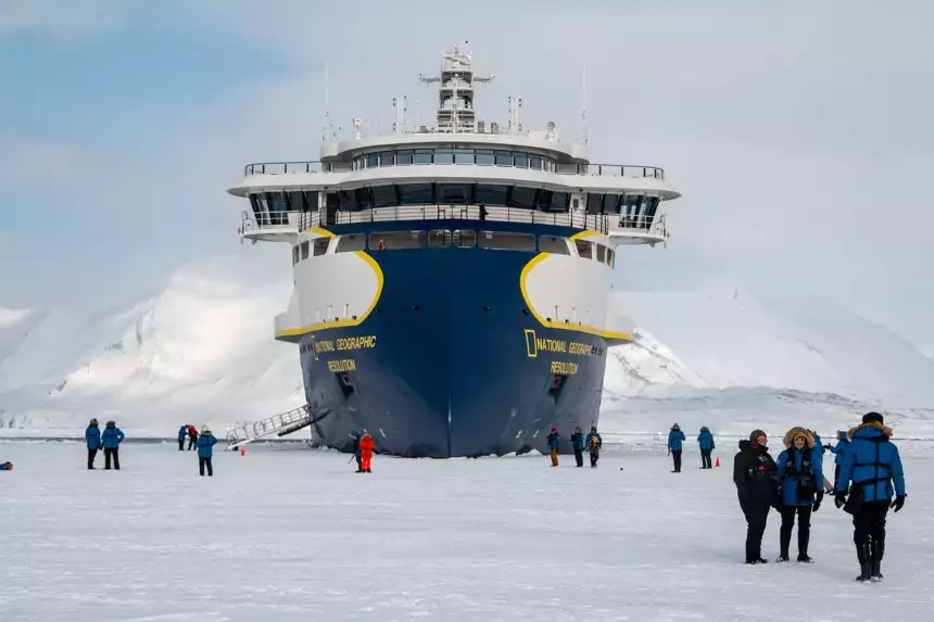 The futuristic blue yellow and white Nat Geo Resolution ship parks in the ice as it's guests walk on the frozen sea around it.