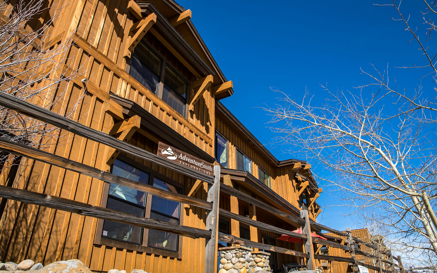 Exterior of AdventureSmith Explorations' office with brown wood walls, large windows, wood fence & stone pillars under sun.