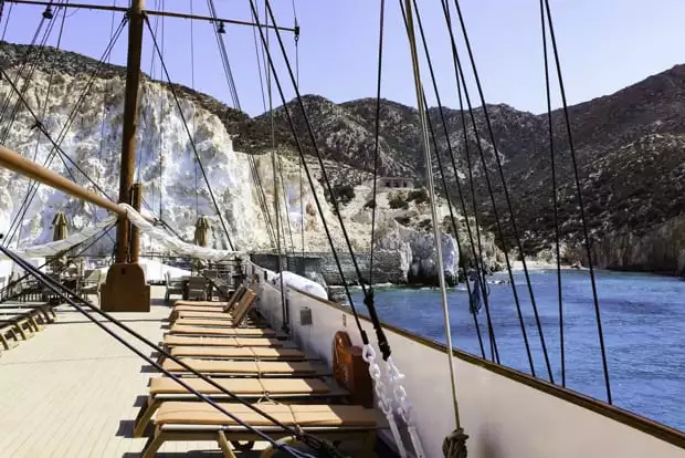 Deck view from the small sail ship the Galileo on the bright blue waters next to a white hillside.