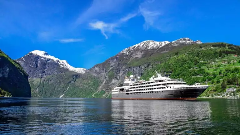 Small expedition ship with gray hull & white upper decks cruises in glassy water by lush green mountains with snowy peaks.