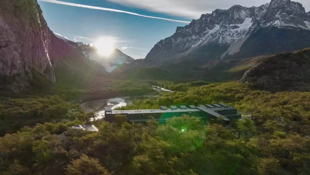 Aerial view of a dark metal, rectangular hotel sitting beside a river in a valley of trees by tall, snow-capped peaks.