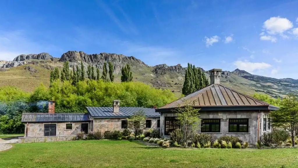 Exterior of stone building with 3 large interconnecting pieces sitting among grass & rugged peaks under a blue sky in Patagonia.