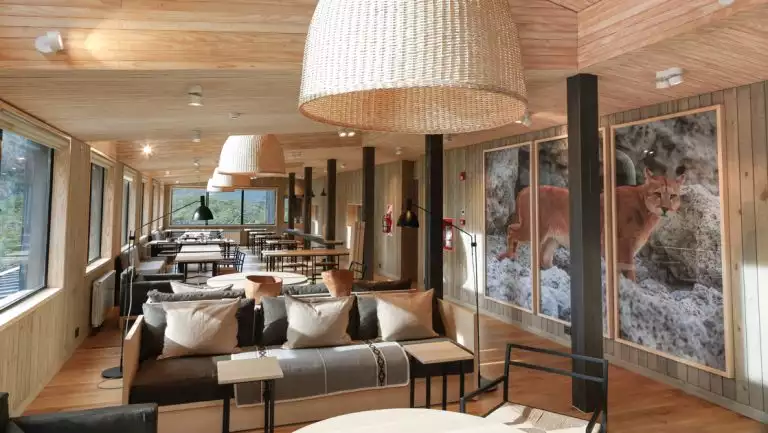 Common area at Hotel Explora El Chalten with light wood, panoramic windows, couches & coffee tables under woven lamps & wildlife art.