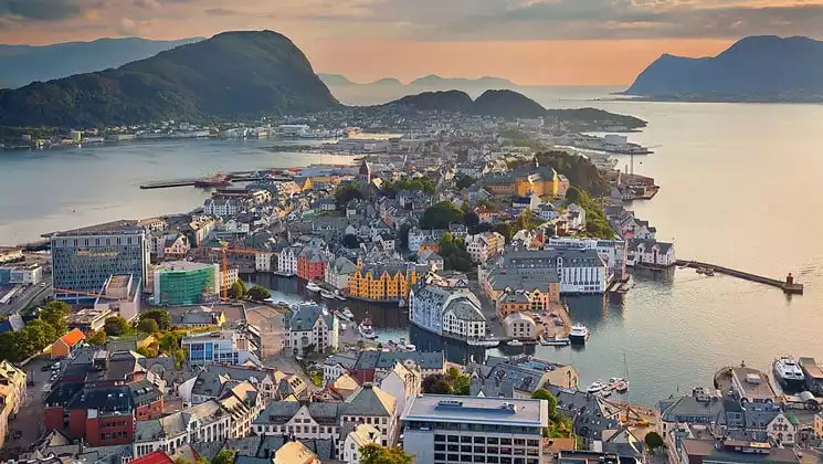Aerial view of water-surrounded small town of various colorful buildings seen at sunset, surrounded by mountains.