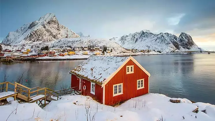 Red house with white trim sits at dusk beside a glassy fjord with mountains across the water & snow everywhere.