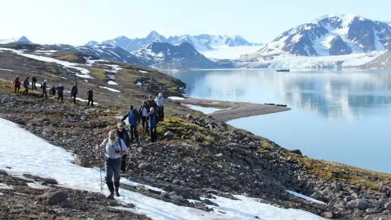 A group of travelers hike toward the camera on a rocky shoreline with the ocean, a ship and mountains behind them in the distance.