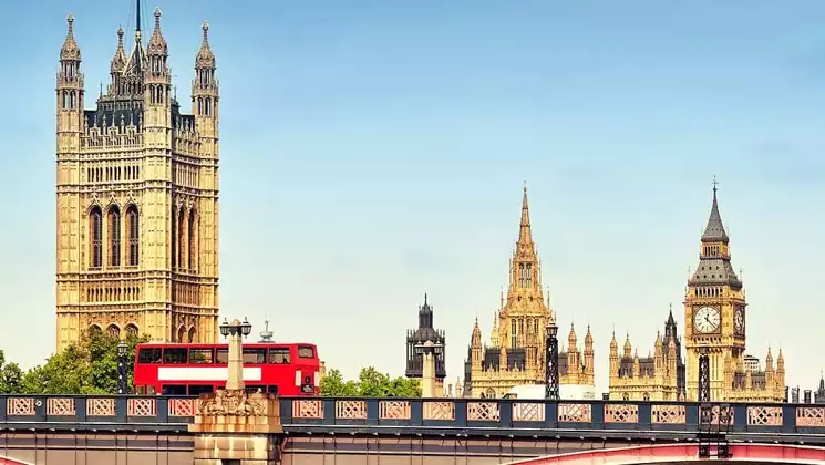 Red double-decker bus drives across a bridge in London with Big Ben & Victoria Tower in the distance on a sunny day.