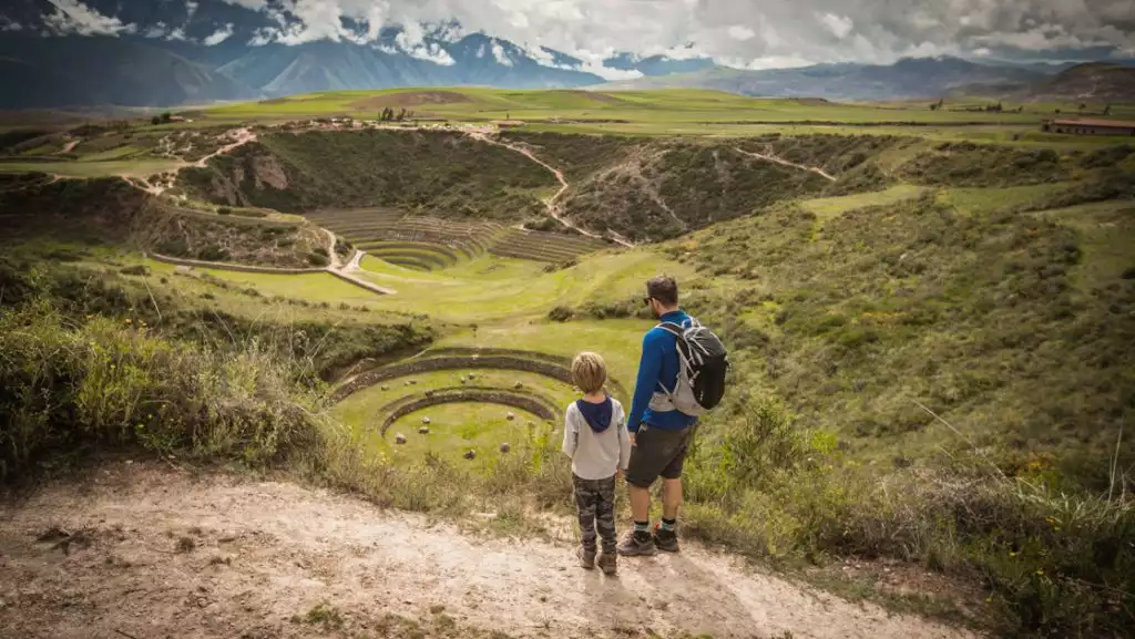 Man & child stand atop a grassy field overlooking a large crater with grassy circles in Sacred Valley, Peru.