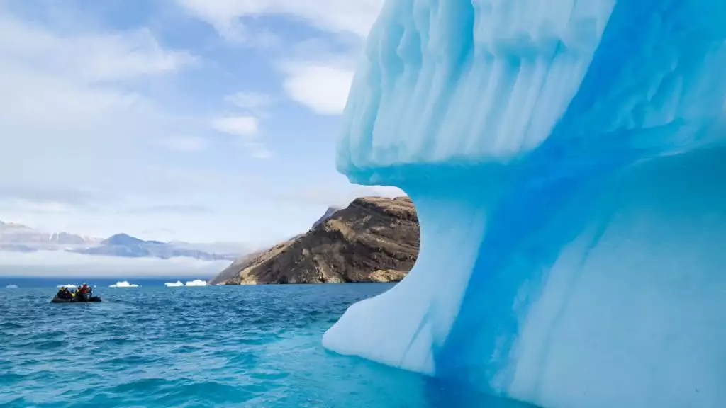 A giant blue iceberg sits on the water in the foreground with a peek of rocky land and a zodiac in the water behind it.