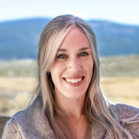 Portrait of Adventure Coordinator Olivia Williams. Blonde female wearing grey sweater leaning on wooden fence