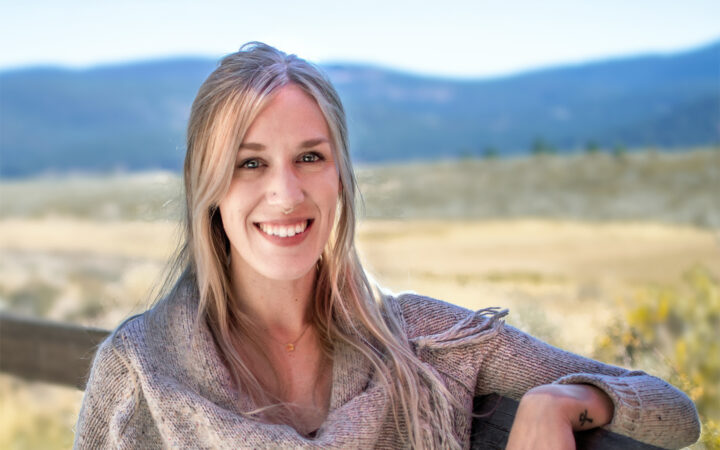 Portrait of Adventure Coordinator Olivia Williams. Blonde female wearing grey sweater leaning on wooden fence