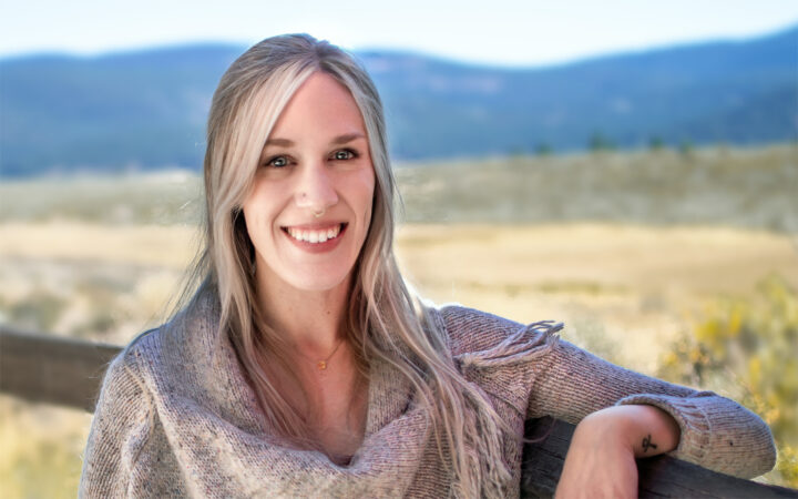 Portrait of Adventure Coordinator Olivia Williams. Blonde female wearing grey sweater leaning on wooden fence