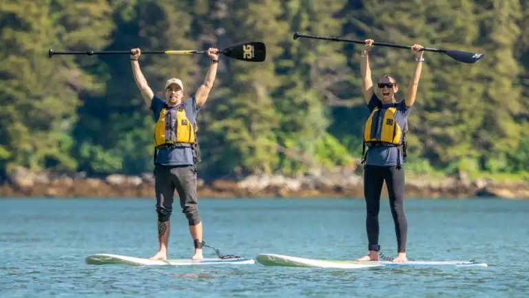 2 paddleboarders raise their paddles above their heads on a sunny day on the Glacier Bay National Park, Haines & Pelican cruise.