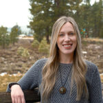 AdventureSmith Olivia Williams standing against a wooden fence next to some pine trees smiling on a cloudy day