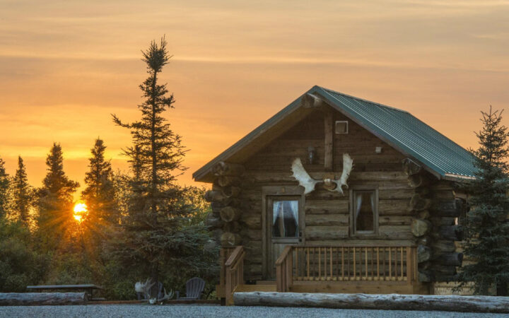 Log cabin with moose antlers above door sits by gravel lot backed by trees in golden sunset at Alaska's Gold Creek Lodge.