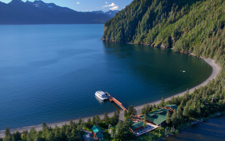 Aerial view of slim peninsula with dock, white boat & small log cabins with green metal roofs beside tall green mountains.