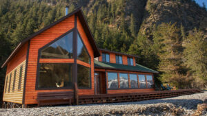 Main lodge of Kenai Fjords Wilderness Lodge with large windows, warm wood exterior, sun deck, & snow-covered mountain views.
