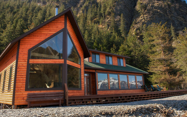 Main lodge of Kenai Fjords Wilderness Lodge with large windows, warm wood exterior, sun deck, & snow-covered mountain views.