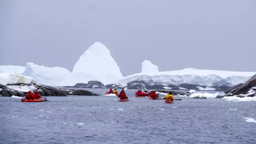 Group of kayakers in red parkas and red double kayaks paddle in the dark waters of Antarctica around icebergs on an overcast day. 