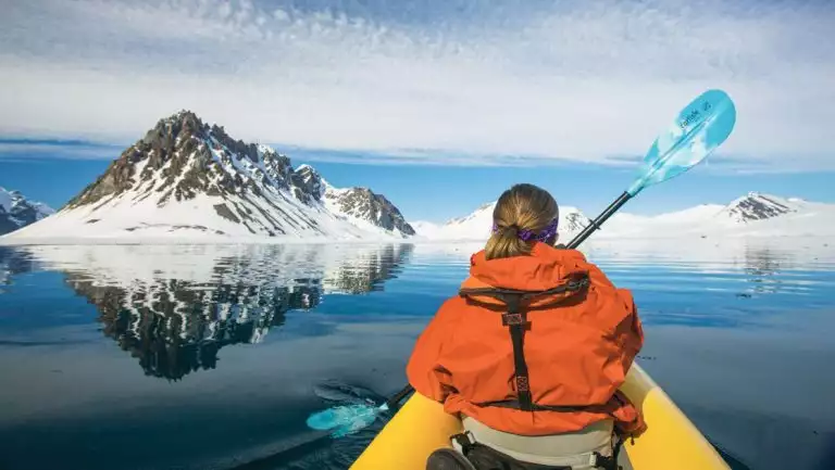 Girl in orange jacket paddles a sit-on-top yellow kayak in glassy water beside snowy mountains on a Norwegian Discovery cruise.