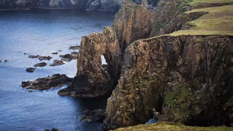 Aerial view of cliffs leading into the sea with one forming an arch & green grass above, seen on an Arctic Saga cruise.