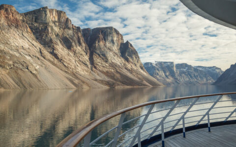 View from empty ship deck with wraparound railing & teak decking, in glassy fjord with tan cliff bands under clouds.