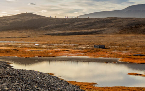 Northwest Passage cruise guests walk a ridgeline on a sloping mountain overlooking autumnal tundra & a small pond.